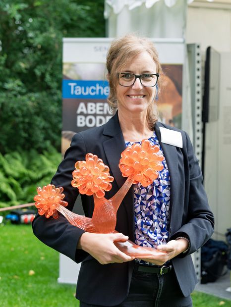 Portrait photograph of Dr Cecilia G. Flocco smiling as she holds an orange, branched model of a myxobacterium in her hands. A banner of the Leibniz Institute DSMZ can be seen in the background.