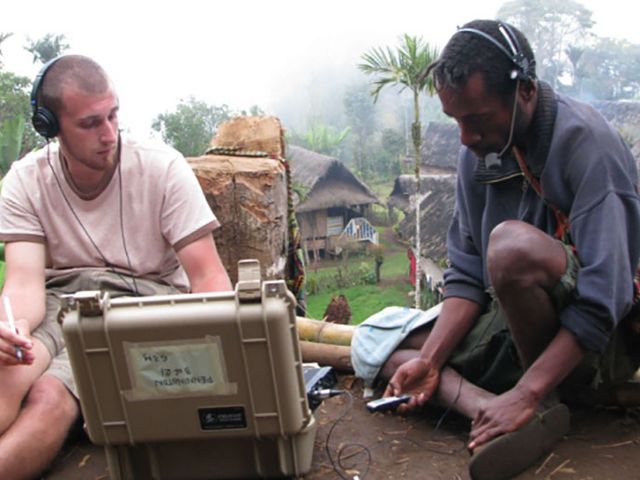 Zwei Männer sitzen auf dem Boden. Beide tragen Kopfhöhrer und beugen sich über ein technisches Gerät (Aufnahmegerät) in einem silbernen Koffer. Im Hintergrund sind der tropische Wald und Hütten zu sehen.