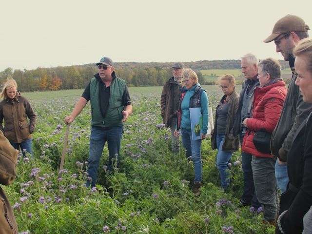 Eine Gruppe von zehn Personen steht auf einem blühenden Feld mit lila Blütenpflanzen. Ein Mann mit Sonnenbrille, dunkler Weste und Basecap hält eine Schaufel  und spricht gestikulierend zu den anderen. Die Gruppe, bestehend aus Männern und Frauen in Outdoor-Kleidung, hört aufmerksam zu. Im Hintergrund sind leicht hügelige Landschaften mit Bäumen und Wiesen zu sehen, die in herbstlichen Farben leuchten